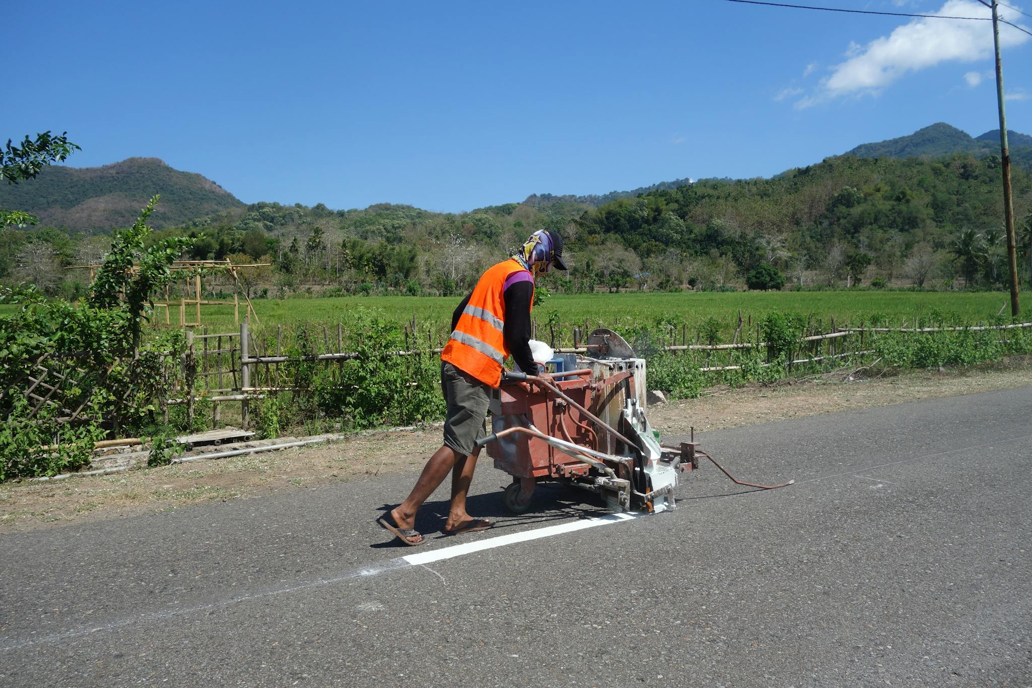 A construction worker uses machinery to paint road lines in a rural area on a sunny day.